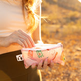 Close-up of a woman opening a Roamio by Bumkins Snack Bag 4 Pack: Disco Boots outdoors in golden sunlight with a desert landscape behind.