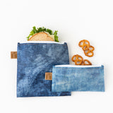 Two bags from Roamio by Bumkins Snack Bag 4 Pack: Denim On Denim rest on a white background, one with a sandwich, the other with pretzels.