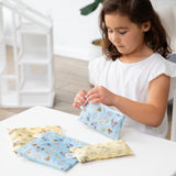 A young girl opens Bumkins Snack Bag 4 Pack: Mickey + Minnie Mouse Brunch Bunch at a white table in a bright playroom.