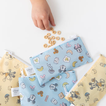 Close-up of a child's hand reaching for cereal near Bumkins Snack Bag 4 Pack: Mickey + Minnie Mouse Brunch Bunch on a white table.