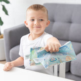 A smiling boy in a white shirt sits at a white table, holding two bags from Bumkins Snack Bag 4 Pack: Stitch Star-ring Up Trouble.