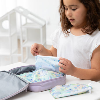A young girl in a white shirt packs Bumkins Snack Bag 4 Pack: Stitch Star-ring Up Trouble in a purple lunch bag on a white table.