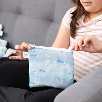 A girl in a striped shirt sits on a gray couch, unzipping a bag from Bumkins Snack Bag 4 Pack: Stitch Star-ring Up Trouble.