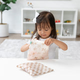 At a white table, a girl with a bow opens a Bumkins Reusable Snack Bag, Large 2 Pack: Palm Check. Shelves are in the background.