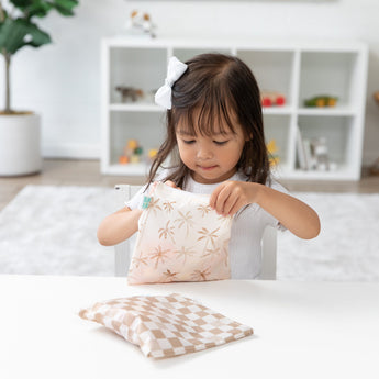 At a white table, a girl with a bow opens a Bumkins Reusable Snack Bag, Large 2 Pack: Palm Check. Shelves are in the background.