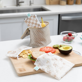 Bags from Bumkins Reusable Snack Bag 4 Pack: Palm Check rest in a lunch bag and on a kitchen counter with a sandwich, fruit, & veggies.