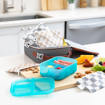 A kitchen counter with Bumkins Lunchbox Legends 6-Piece Set: Double Check Blue Jelly, lunch bag, cutting board, and fruit & veggies.