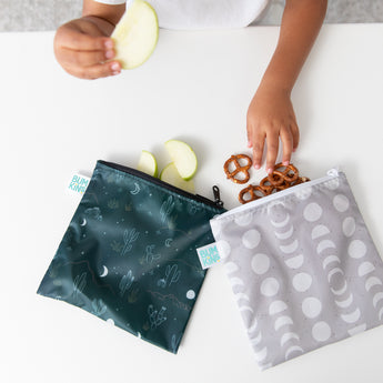 A child's hands hold apple slices as they reach for pretzels from Bumkins Reusable Snack Bag, Large 2 Pack: Desert Night on a white table.