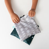 A child holds Bumkins Reusable Snack Bag, Large 2 Pack: Desert Night on a white table.