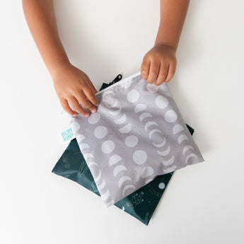 A child holds Bumkins Reusable Snack Bag, Large 2 Pack: Desert Night on a white table.
