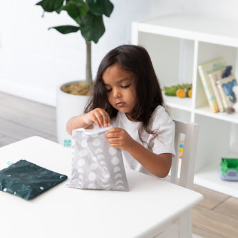 A young child opens a Bumkins Reusable Snack Bag 4 Pack: Desert Night, with another bag and books in the background.