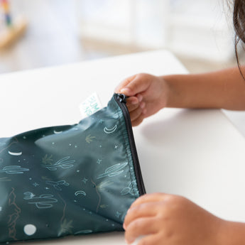 A child zips a Bumkins Reusable Snack Bag, Large 2 Pack: Desert Night at a white table.