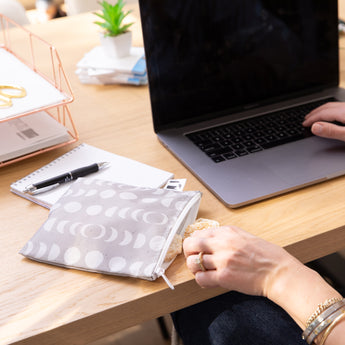 A hand reaches for snacks from a Roamio by Bumkins Reusable Snack Bag 4 Pack: Desert Night on a desk by a notepad, pen, and laptop.