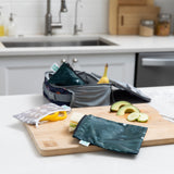 A Bumkins Reusable Snack Bag, Large 2 Pack: Desert Night holds a sandwich next to cucumber and avocado on a cutting board on a kitchen counter.