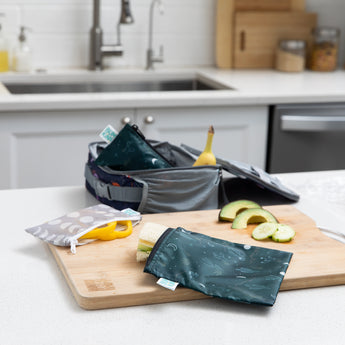 A Bumkins Reusable Snack Bag, Large 2 Pack: Desert Night holds a sandwich next to cucumber and avocado on a cutting board on a kitchen counter.