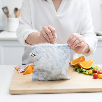 A person zips a bag from Bumkins Snack In Style 3-Piece Set: Disco Boots on a cutting board with fruit and veggies.