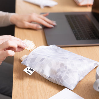 A person using a laptop grabs a rice cake from a Roamio by Bumkins Reusable Snack Bag, Large 2 Pack: Disco Boots nearby on a desk.