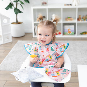 A girl seated in a high chair enjoys snacks with Bumkins Snack In Style 3-Piece Set: Disco Boots in a bright playroom.