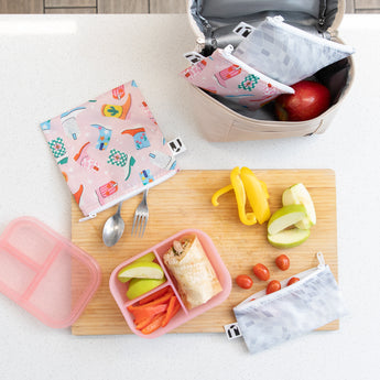 Aerial view of a kitchen counter with Bumkins Lunchbox Legends 6-Piece Set: Disco Pink Jelly, lunch bag, cutting board, and fruit & veggies.