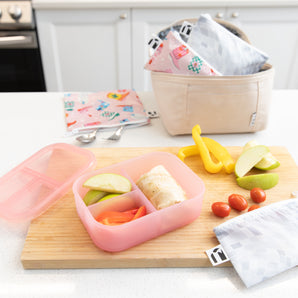A kitchen counter with Bumkins Lunchbox Legends 6-Piece Set: Disco Pink Jelly, lunch bag, cutting board, and fruit & veggies.