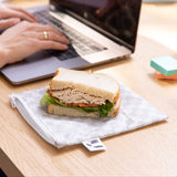 A sandwich sits on top of a Roamio by Bumkins Snack Bag 4 Pack: Rich & Boujee on a wood desk next to a person typing on a laptop.