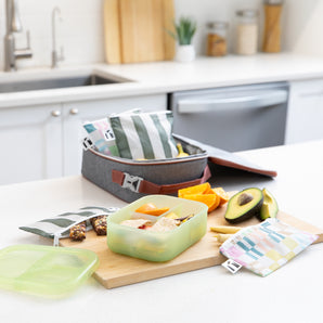 A kitchen counter with Bumkins Lunchbox Legends 6-Piece Set: Motley Green Jelly, lunch bag, cutting board, and fruit & veggies.