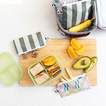 A kitchen counter with Bumkins Lunchbox Legends 6-Piece Set: Motley Green Jelly, lunch bag, cutting board, and fruit & veggies.