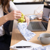 A person places grapes in a bag from Bumkins Lunchbox Legends 6-Piece Set: Coastal Purple Jelly on a desk by a laptop & notebook.