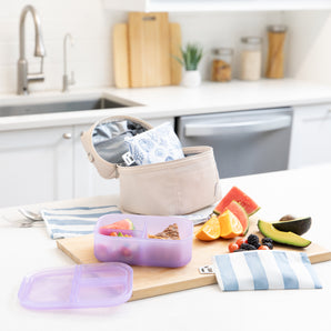 A kitchen counter with Bumkins Lunchbox Legends 6-Piece Set: Coastal Purple Jelly, lunch bag, cutting board, and fruit & veggies.