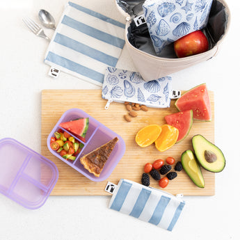 A kitchen counter with Bumkins Lunchbox Legends 6-Piece Set: Coastal Purple Jelly, lunch bag, cutting board, and fruit & veggies.