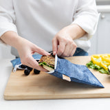 A person places a sandwich in a bag from Bumkins Snack In Style 3-Piece Set: Denim On Denim on a cutting board with fruit and veggies.