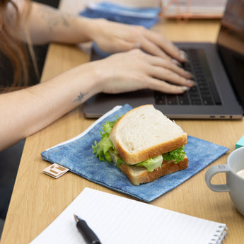 A sandwich sits on top of a Roamio by Bumkins Reusable Snack Bag, Large 2 Pack: Denim on Denim on a desk next to a person using a laptop.