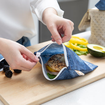 A person opens a Roamio by Bumkins Reusable Snack Bag, Large 2 Pack: Denim on Denim on a cutting board revealing a sandwich inside.