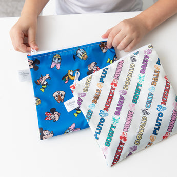 A child holds Bumkins Reusable Snack Bag, Large 2 Pack: Mickey & Friends MVP on a white table.