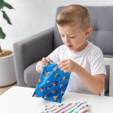 A boy in a white shirt opens Bumkins Reusable Snack Bag, Large 2 Pack: Mickey & Friends MVP at a table in front of a couch and potted plant.