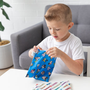A boy in a white shirt opens Bumkins Reusable Snack Bag, Large 2 Pack: Mickey & Friends MVP at a table in front of a couch and potted plant.
