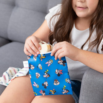 A girl removes a sandwich from Bumkins Reusable Snack Bag, Large 2 Pack: Mickey & Friends MVP while on the couch.