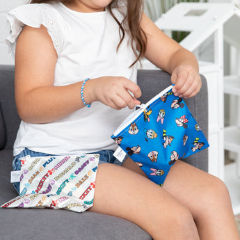 A child wearing a white shirt on a couch zips Bumkins Reusable Snack Bag, Large 2 Pack: Mickey & Friends MVP.