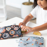 A child reaches for a sandwich in a bag from Bumkins Reusable Snack Bag 4 Pack: Everyday Is Halloween on a table with another bag nearby.