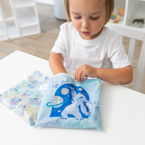 Young child in a white shirt reaches into a Bumkins Snack Bag from the Disney Stitch Universe 4 Pack at a white table.
