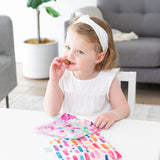 A girl eats snacks and holds bags from Bumkins Lunchbox Legends 6-Piece Set: Watercolor Pink Jelly at a white table in a living room.