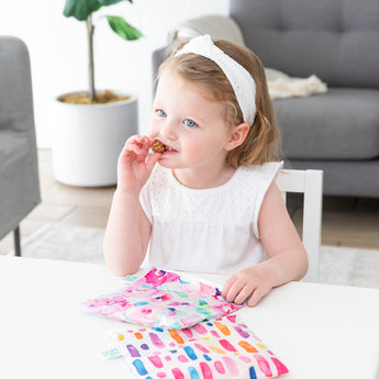 A girl eats snacks and holds bags from Bumkins Lunchbox Legends 6-Piece Set: Watercolor Pink Jelly at a white table in a living room.