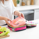 A woman packs Bumkins Lunchbox Legends 6-Piece Set: Watercolor Pink Jelly into an open lunch bag on a kitchen counter by fruit & veggies.