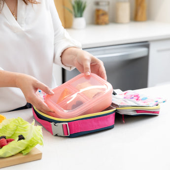 A woman packs Bumkins Lunchbox Legends 6-Piece Set: Watercolor Pink Jelly into an open lunch bag on a kitchen counter by fruit & veggies.