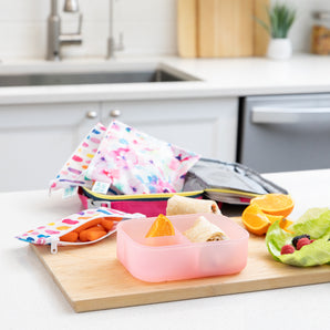 A kitchen counter with Bumkins Lunchbox Legends 6-Piece Set: Watercolor Pink Jelly, lunch bag, cutting board, and fruit & veggies.