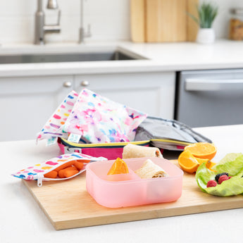 A kitchen counter with Bumkins Lunchbox Legends 6-Piece Set: Watercolor Pink Jelly, lunch bag, cutting board, and fruit & veggies.