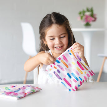 A young girl with long hair smiles while holding a colorful patterned pouch at a table. A chair and table with a plant in the background are slightly blurred.