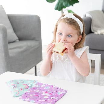 A young girl eats a sandwich with bags from Bumkins Snack In Style 3-Piece Set: Unicorns and Rainbows on a table nearby.