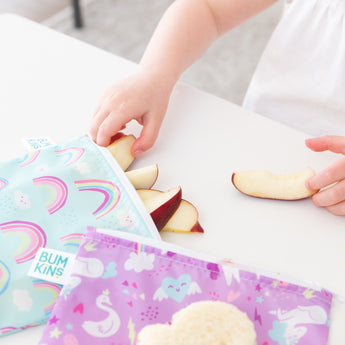Close-up of a child reaching for apple slices from bags from Bumkins Snack In Style 3-Piece Set: Unicorns and Rainbows.