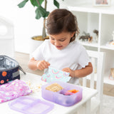 A girl eats lunch with Bumkins Lunchbox Legends 6-Piece Set: Unicorns Purple Jelly near a lunch bag at a white table in a playroom.